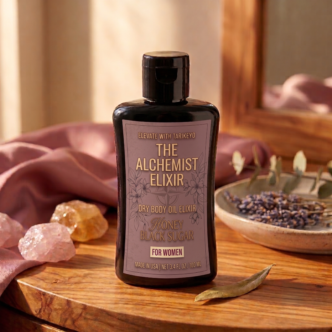 A bottle of Tarkney 'The Alchemist Elixir' dry body oil elixir, set on a rustic wooden table with pink fabric, crystals, and dried lavender in a warm, sunlit environment.